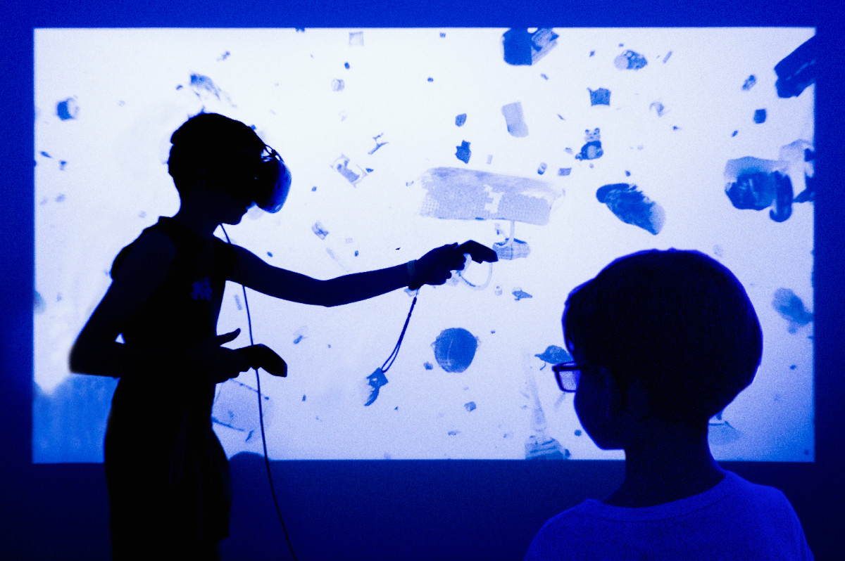 a boy wearing virtual reality headset in front of a blue projected video another boy watching him